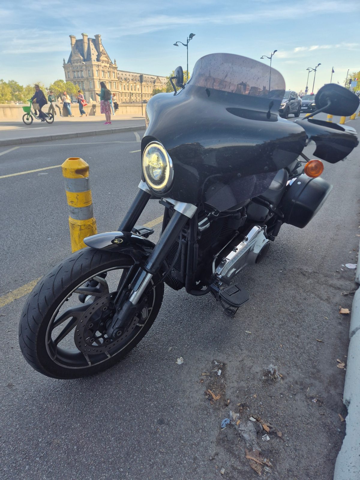 A black Harley-Davidson motorcycle parked kerbside with the Louvre palace and pedestrians in the background in Paris.