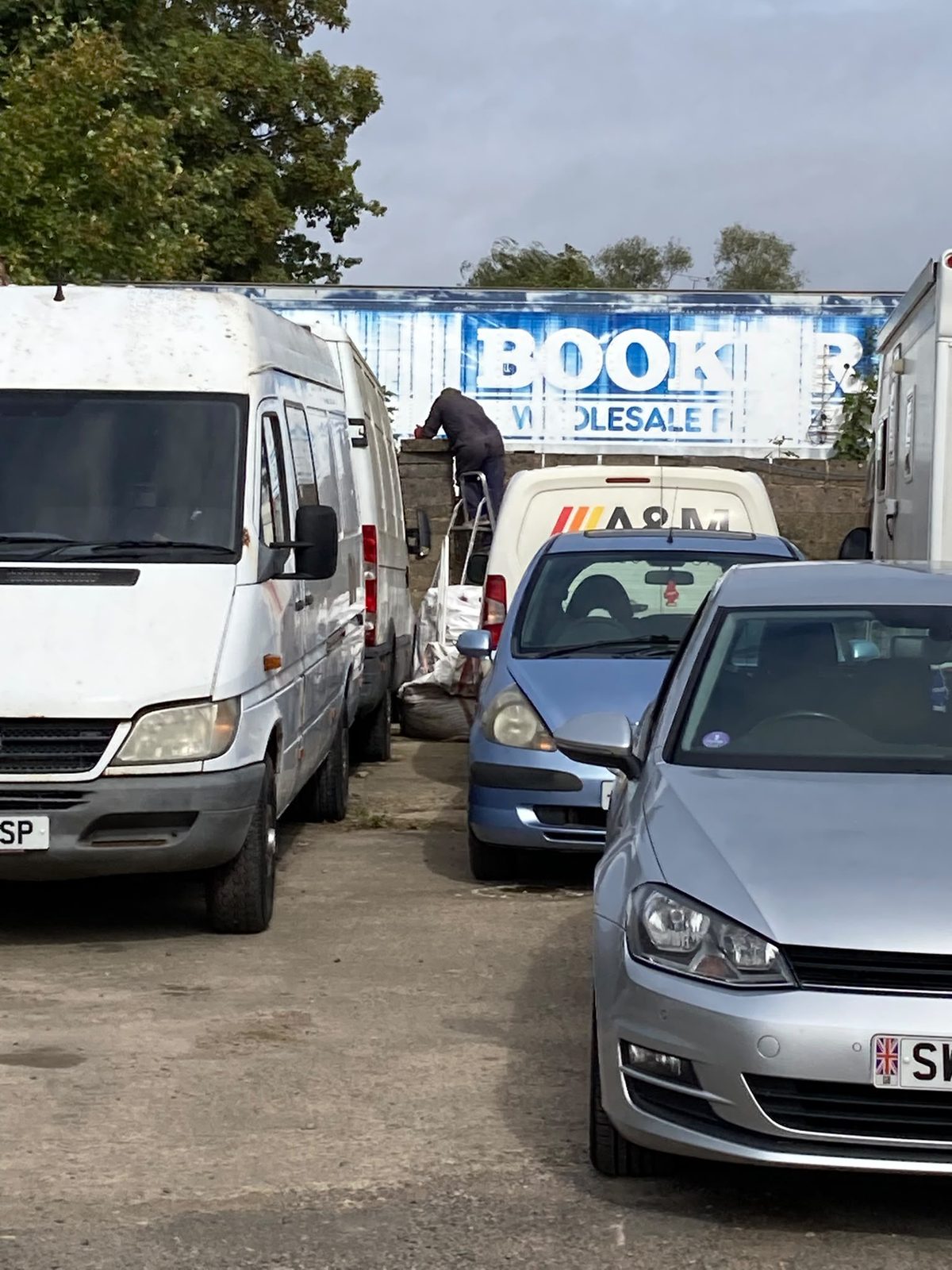 A full garage yard with vans and cars parked close together, and a man on a stepladder working at a wall by a Booker lorry.