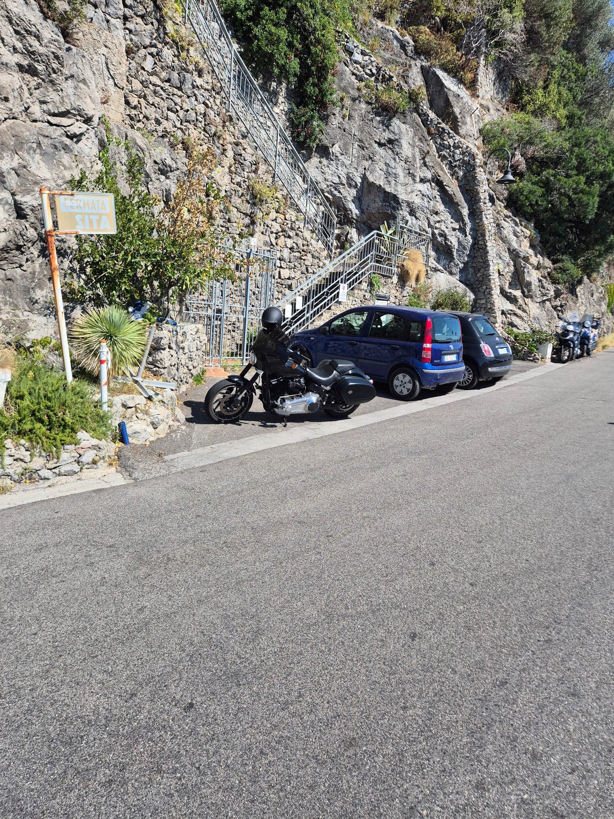 A black Harley-Davidson parked beside a bus stop on a narrow coastal road, next to two small cars with rock cliffs behind.