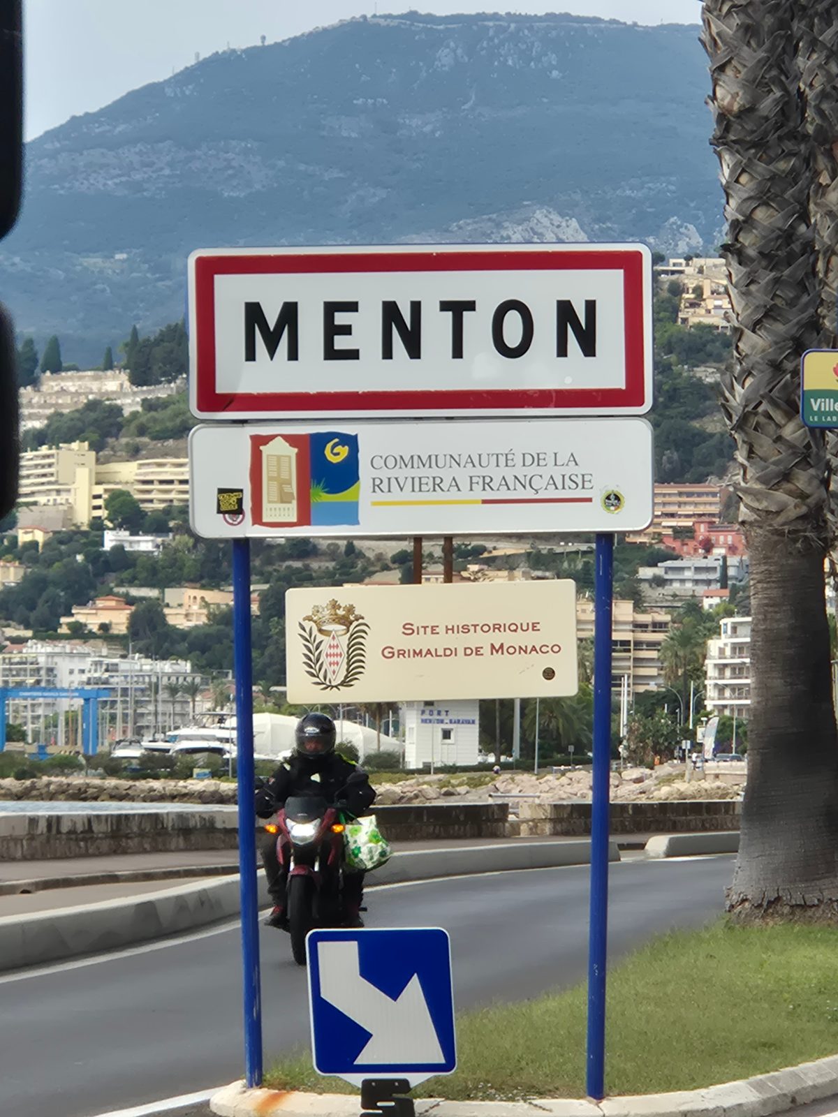 The Menton town sign on the French Riviera with a motorcyclist riding past and hills behind.