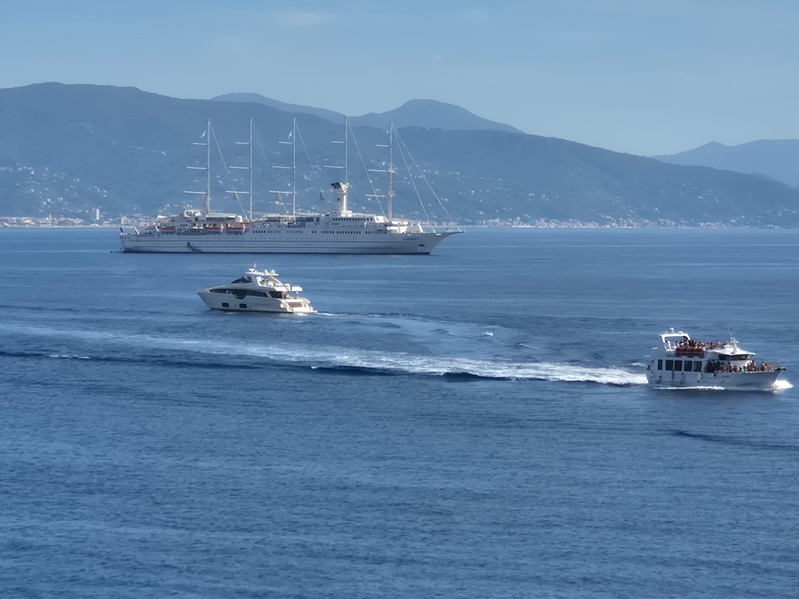 A large white sailing cruise ship anchored in a bay with a motor yacht and a smaller tour boat passing by, mountains in the background.