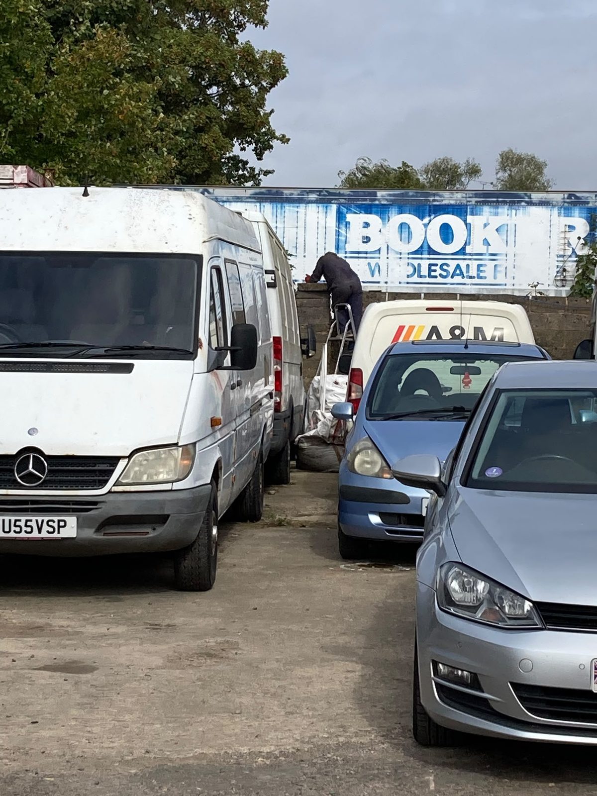 A busy garage yard with vans and cars parked tightly together, and a person on a stepladder working by the boundary wall.