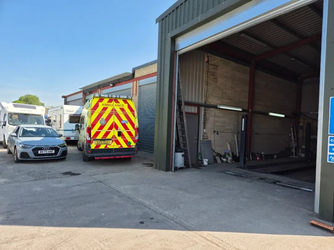 An NHS ambulance van with yellow and green markings parked beside a silver Audi, a motorhome and campervans outside the open roller door of Redvers Services garage.
