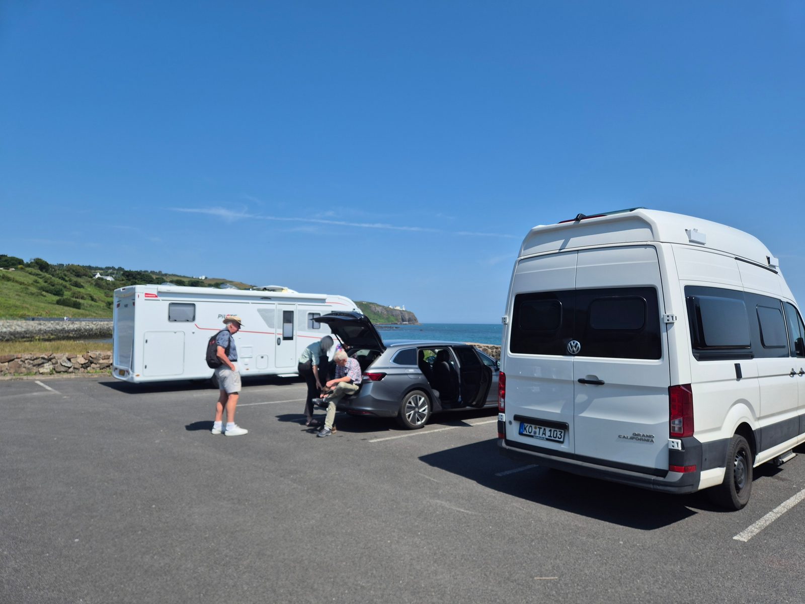 A white Volkswagen Grand California campervan with German plates and a Pilote coachbuilt motorhome parked in a seafront car park, with a silver estate car and three people between them under a clear blue sky.