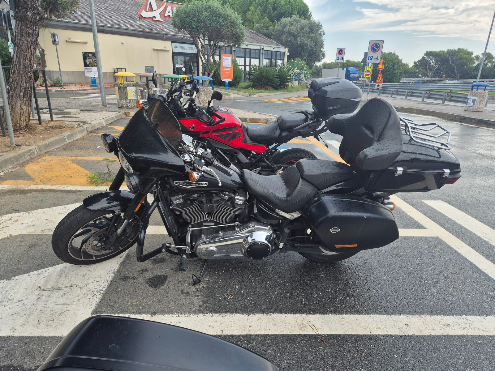 A black Harley-Davidson with panniers and a top box parked beside a red sports motorcycle at a service station car park.