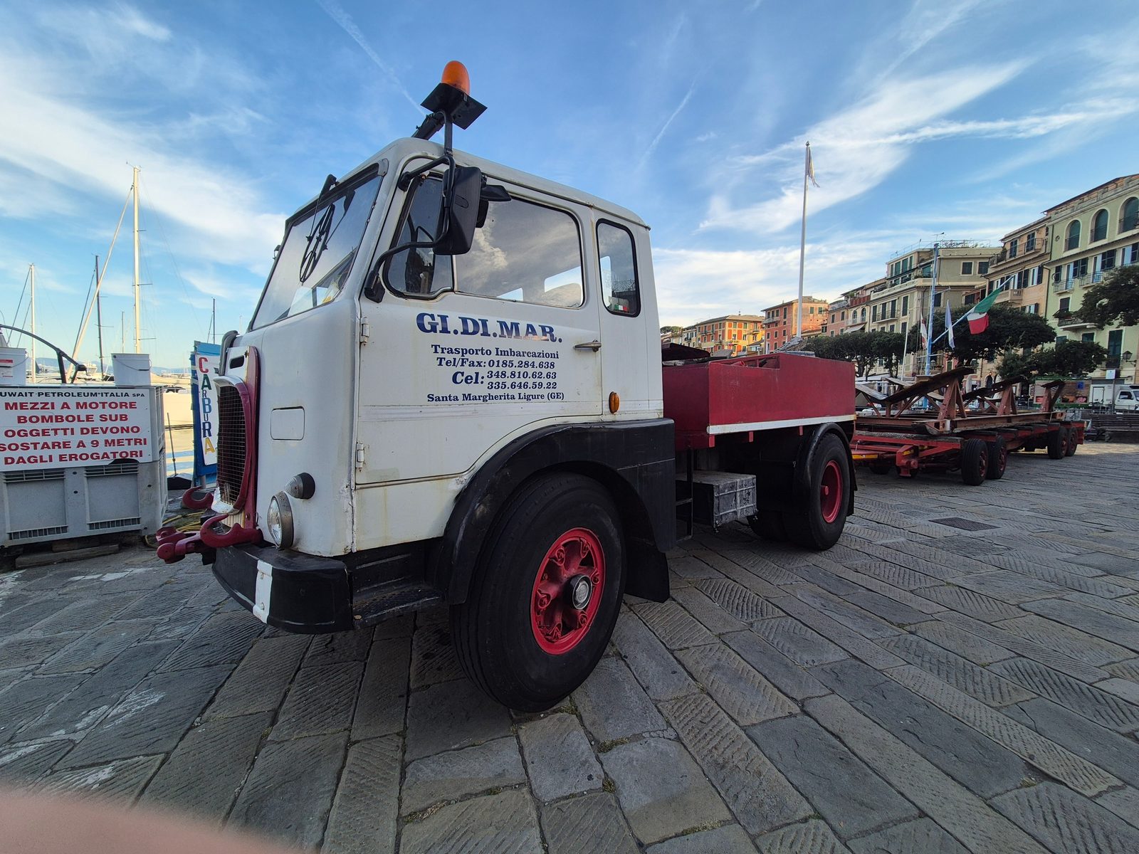 A vintage white Fiat flatbed truck parked on a cobbled quayside with yachts and colourful buildings behind.