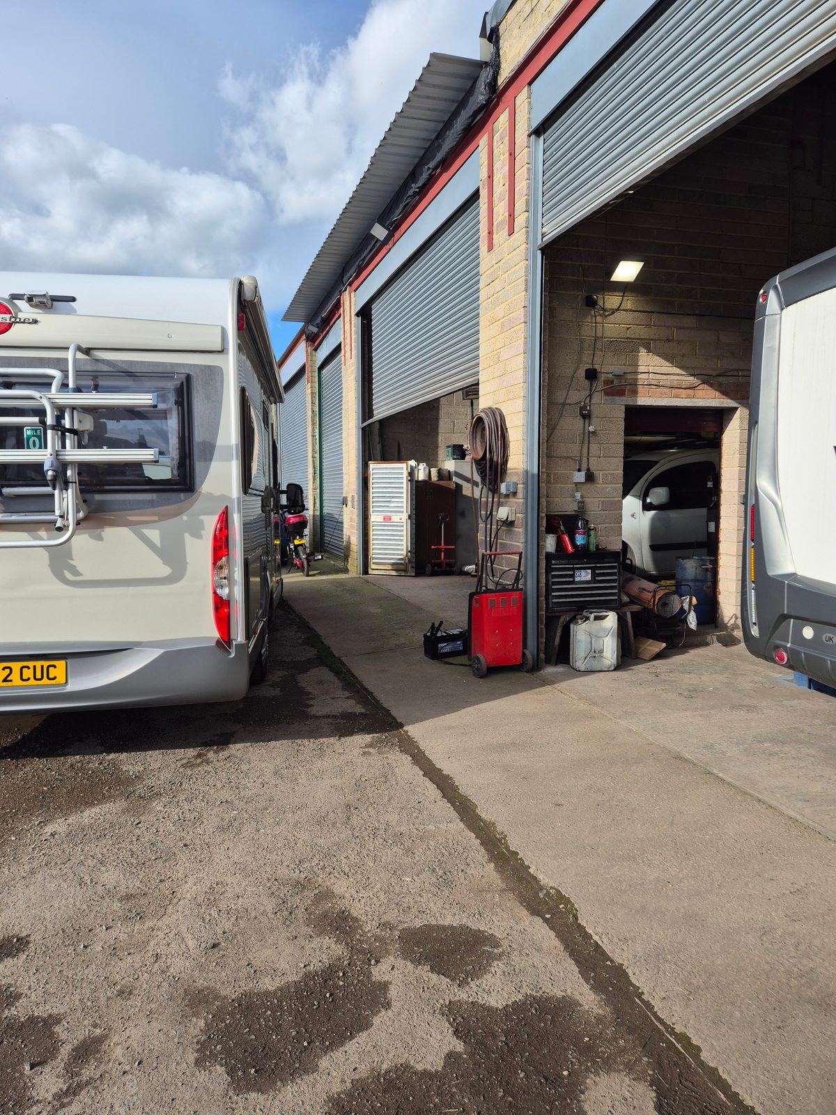 The rear of a coachbuilt motorhome parked beside a garage unit with an open roller shutter, a van inside the bay, and a battery charger, tool chest and jerry cans by the entrance.
