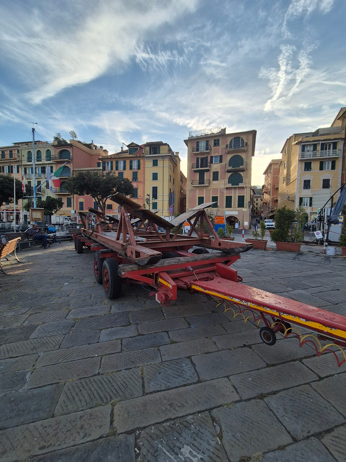 A large red boat trailer parked on a cobbled square surrounded by colourful Italian buildings under a blue sky.