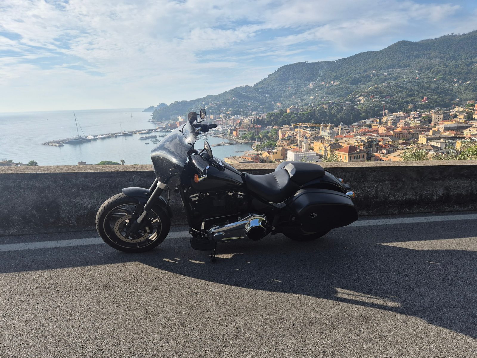 A black Harley-Davidson motorcycle parked by a low wall overlooking an Italian coastal town and harbour.