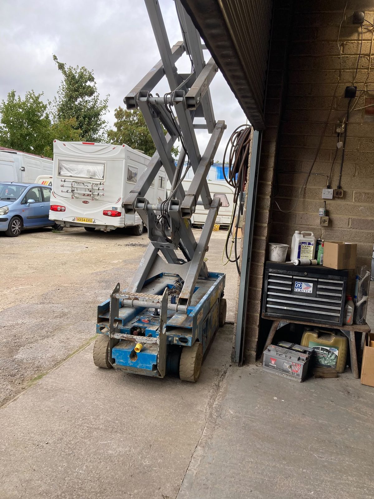 A blue scissor lift parked by an open garage door with a caravan and cars in the yard behind.