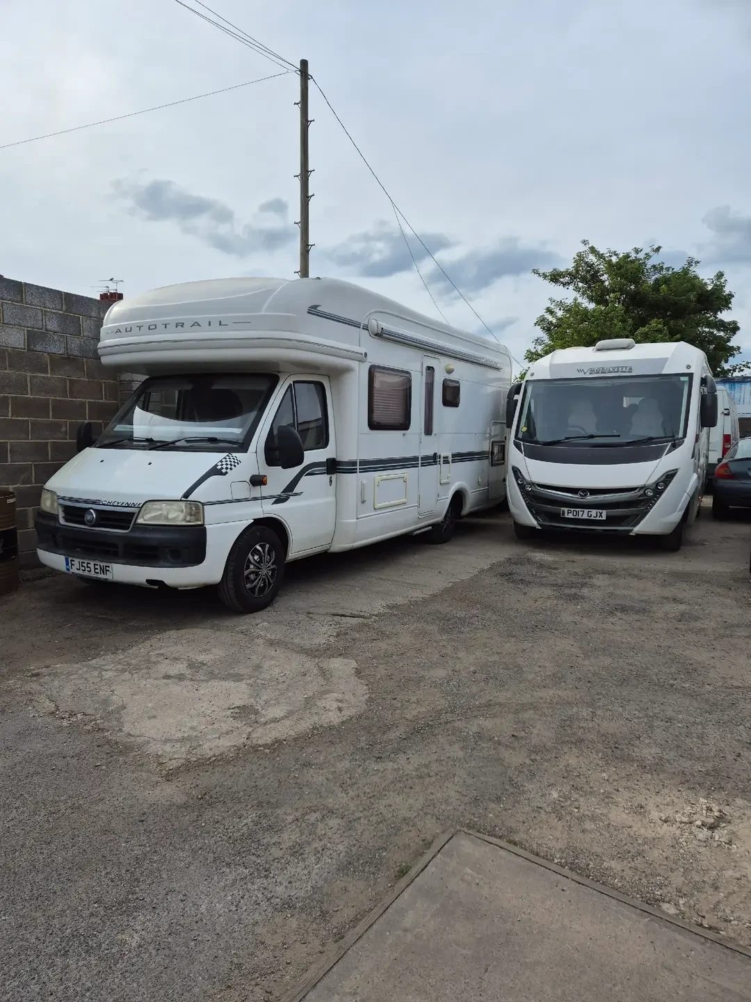 A white Auto-Trail Cheyenne coachbuilt motorhome and a white Niesmann Bischoff motorhome parked side by side on a tarmac yard beside a breeze-block wall.
