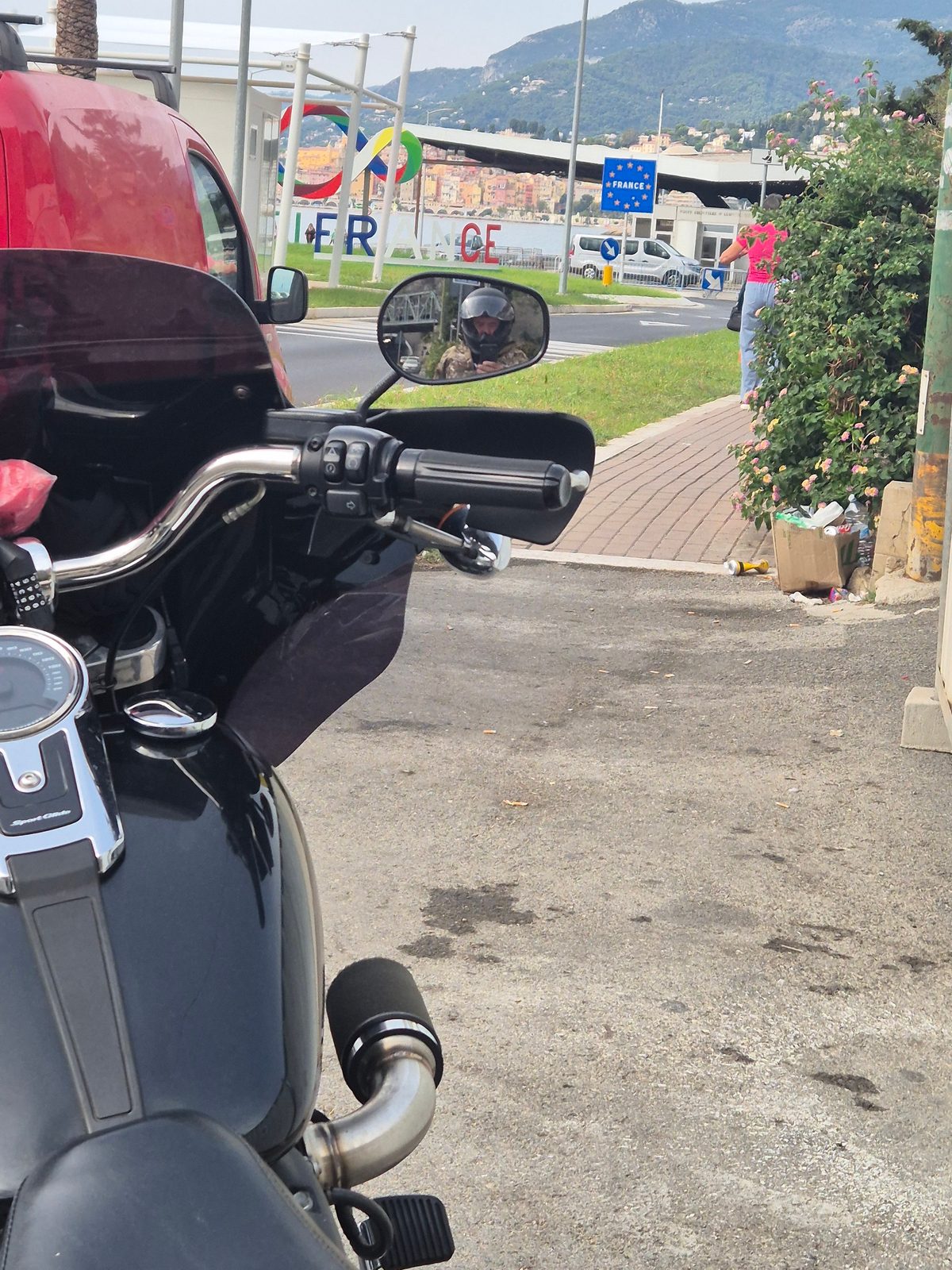 A motorcycle parked near the France border crossing on the Mediterranean coast, with the rider reflected in the mirror.