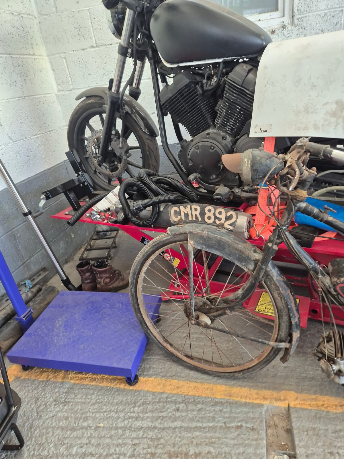 A modern black motorcycle on an MOT ramp beside a very old rusty motorbike with a CMR 892 registration plate.