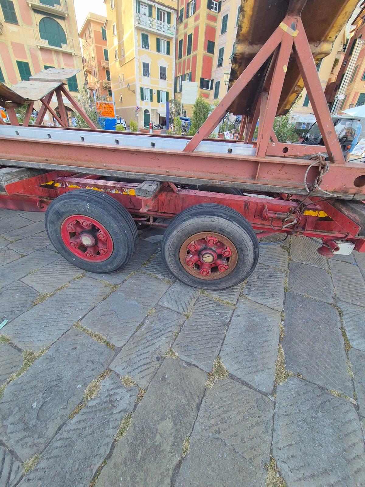 An old red twin-axle trailer parked on stone cobbles with colourful Italian buildings in the background.