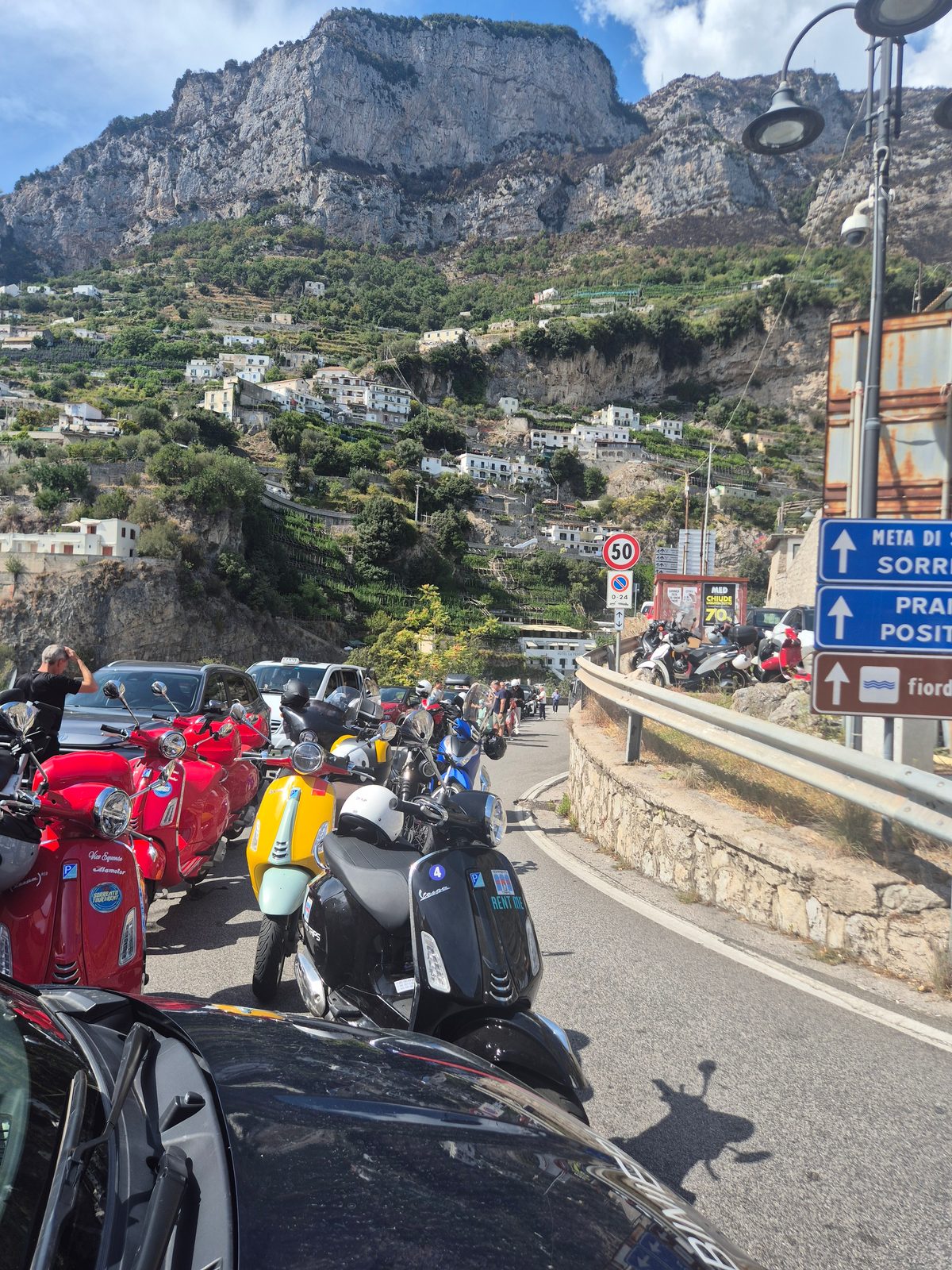 A row of parked Vespa scooters on a roadside in a coastal Italian town with steep cliffs and hillside houses behind.