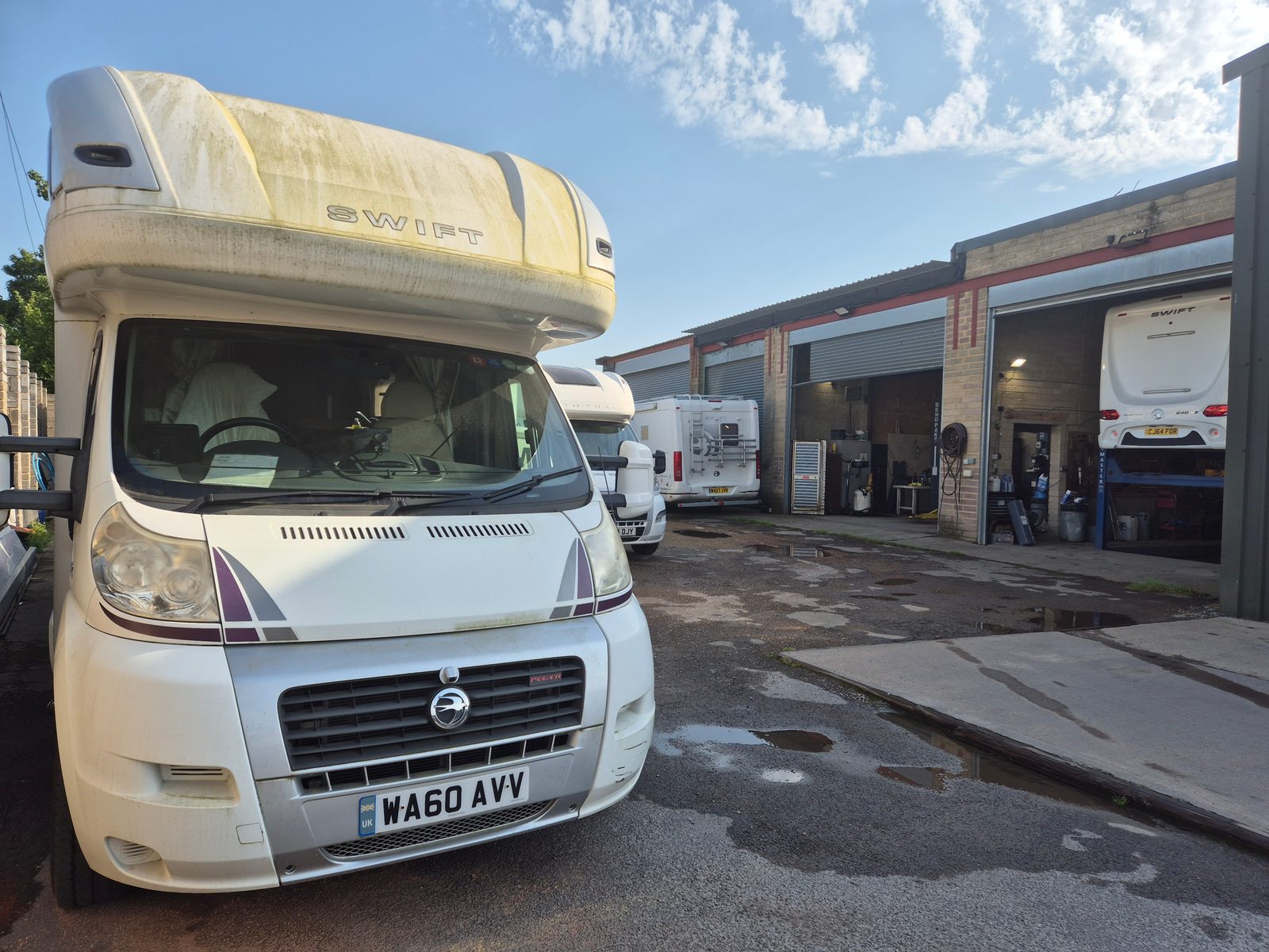 A white Swift coachbuilt motorhome with an overcab bed parked on a yard, with further motorhomes and open workshop bays behind under a blue sky.