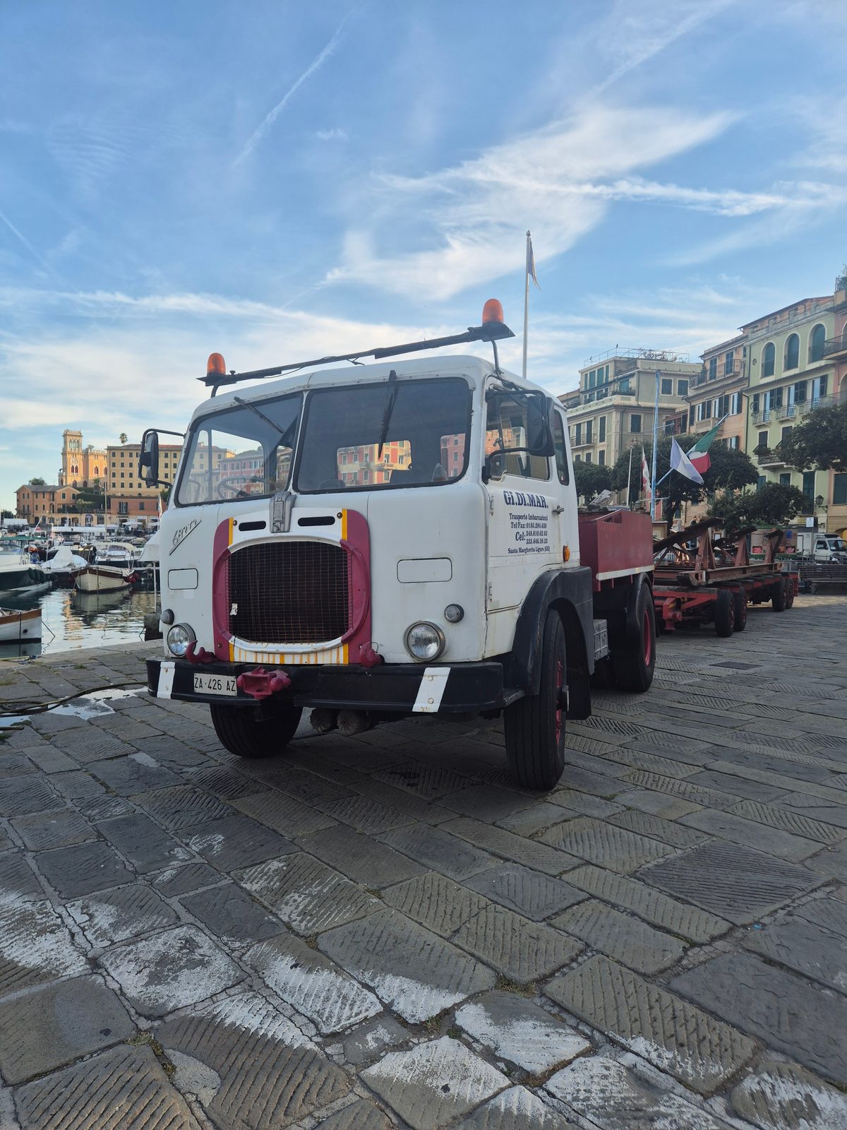 A vintage white and red Fiat truck with a flatbed trailer parked on cobblestones beside a harbour with boats and colourful buildings.