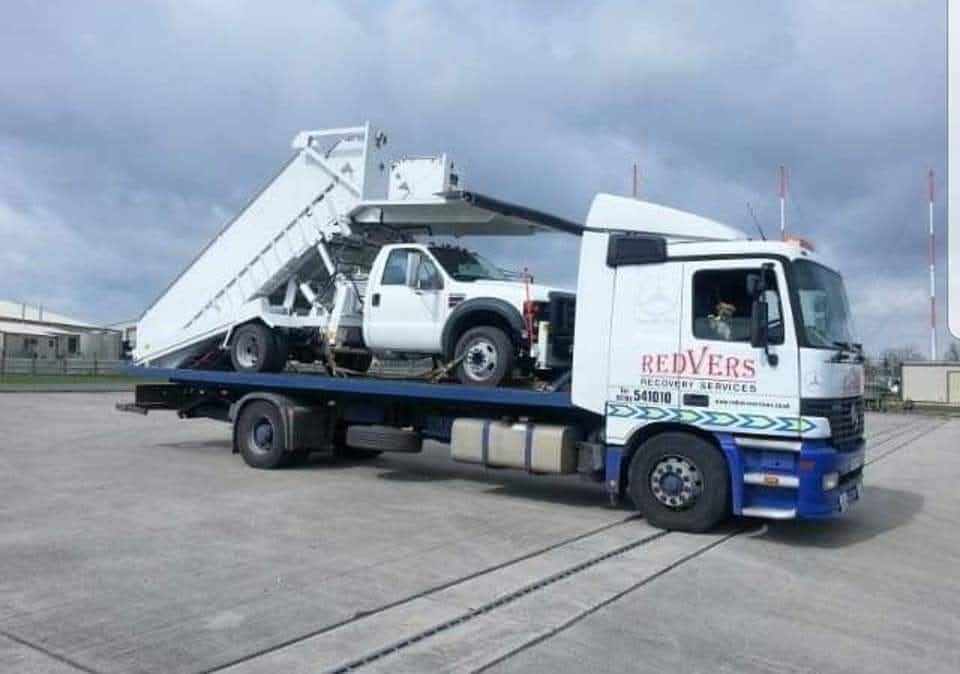 A blue and white Mercedes Redvers Recovery Services flatbed lorry loaded with a white tipper truck on a concrete yard under a cloudy sky.
