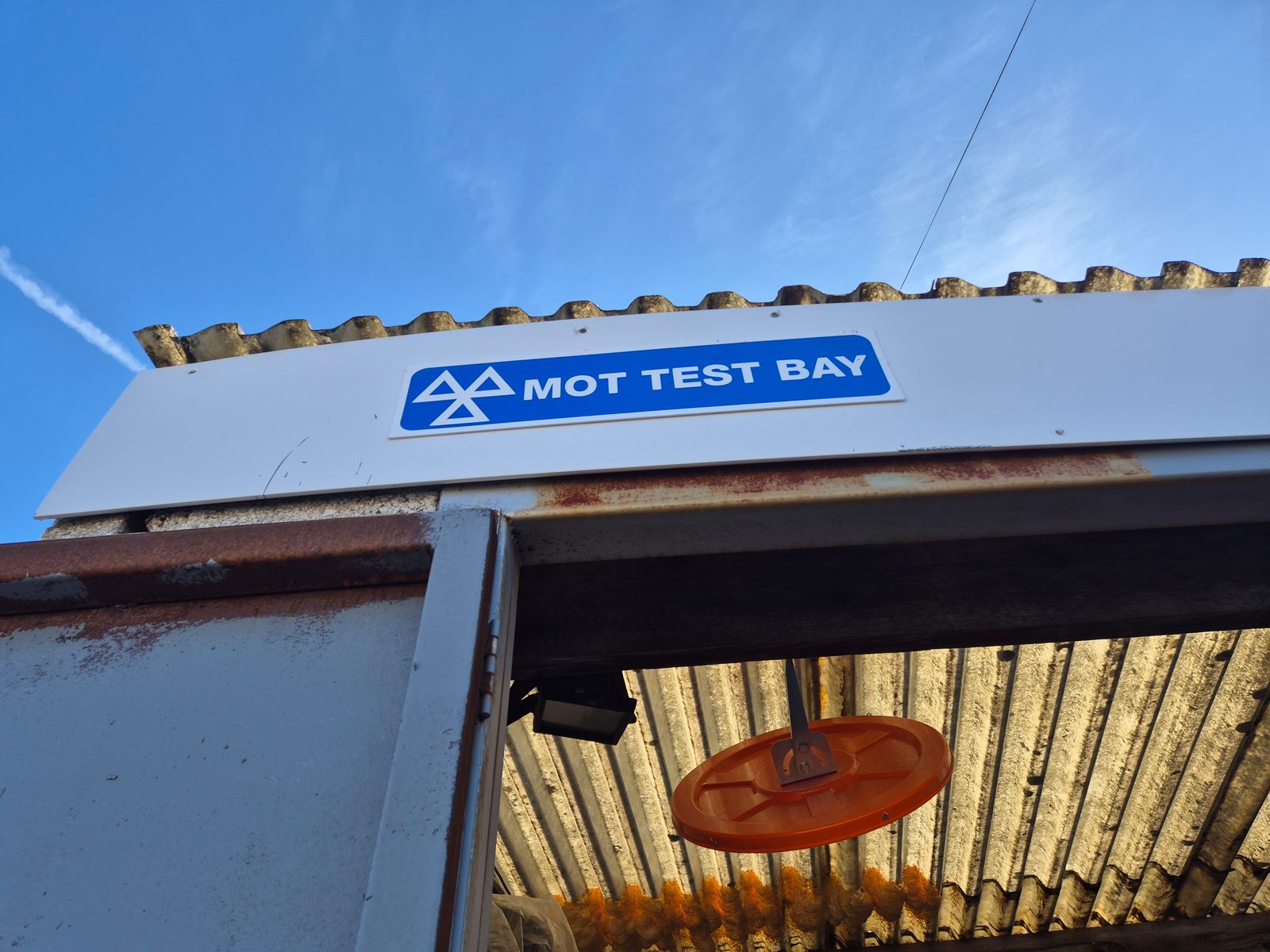 An MOT Test Bay sign above a garage entrance against a bright blue sky.