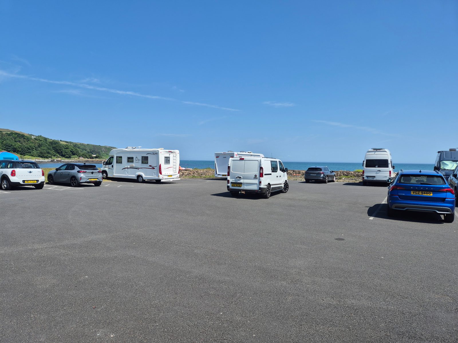 A coastal tarmac car park with a white motorhome, a white panel van, a VW campervan, and several cars parked beside the sea under a clear blue sky.