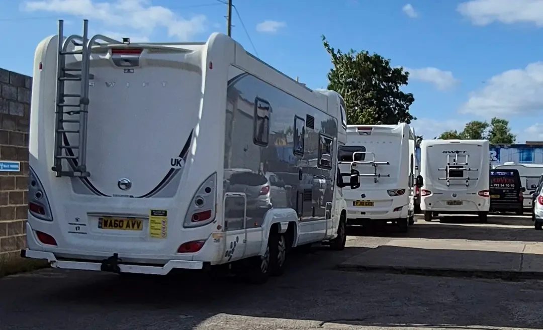 A white Swift coachbuilt motorhome with a rear ladder parked beside other motorhomes and a van in an outdoor forecourt under a blue sky.