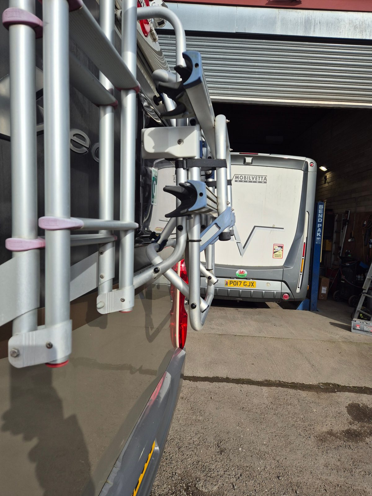View past a rear-mounted ladder and bike rack towards a silver Mobilvetta motorhome raised on a BendPak ramp inside a garage workshop.