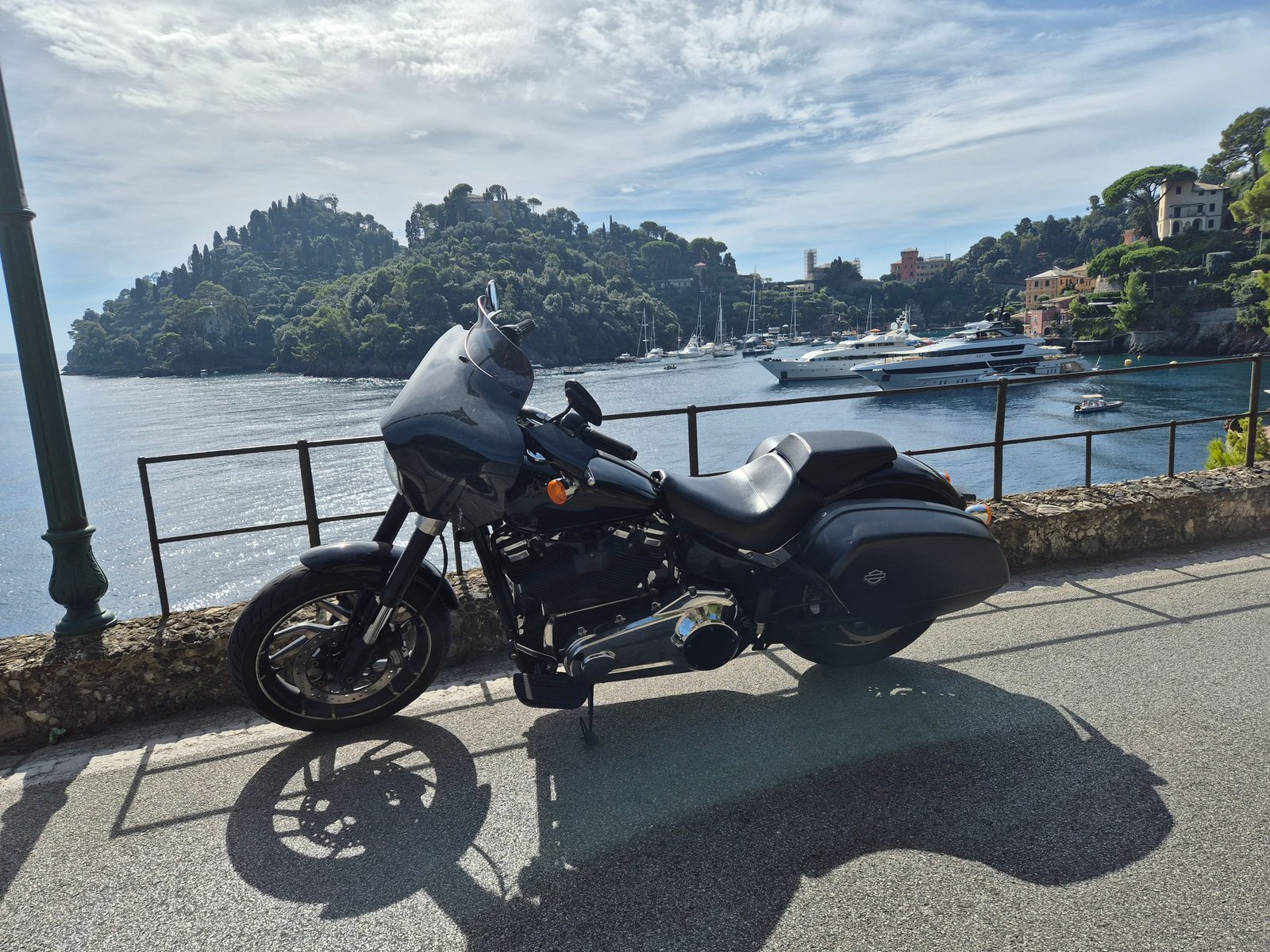 A black Harley-Davidson motorcycle parked by a railing above a harbour full of yachts, with a wooded headland and coastal town behind.
