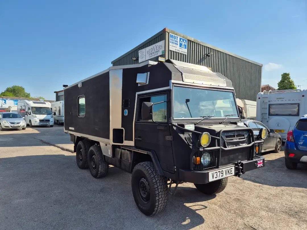 A black six-wheel-drive Pinzgauer-based expedition camper with off-road tyres parked on the forecourt outside the Redvers MOT Testing Station, with motorhomes and cars nearby.