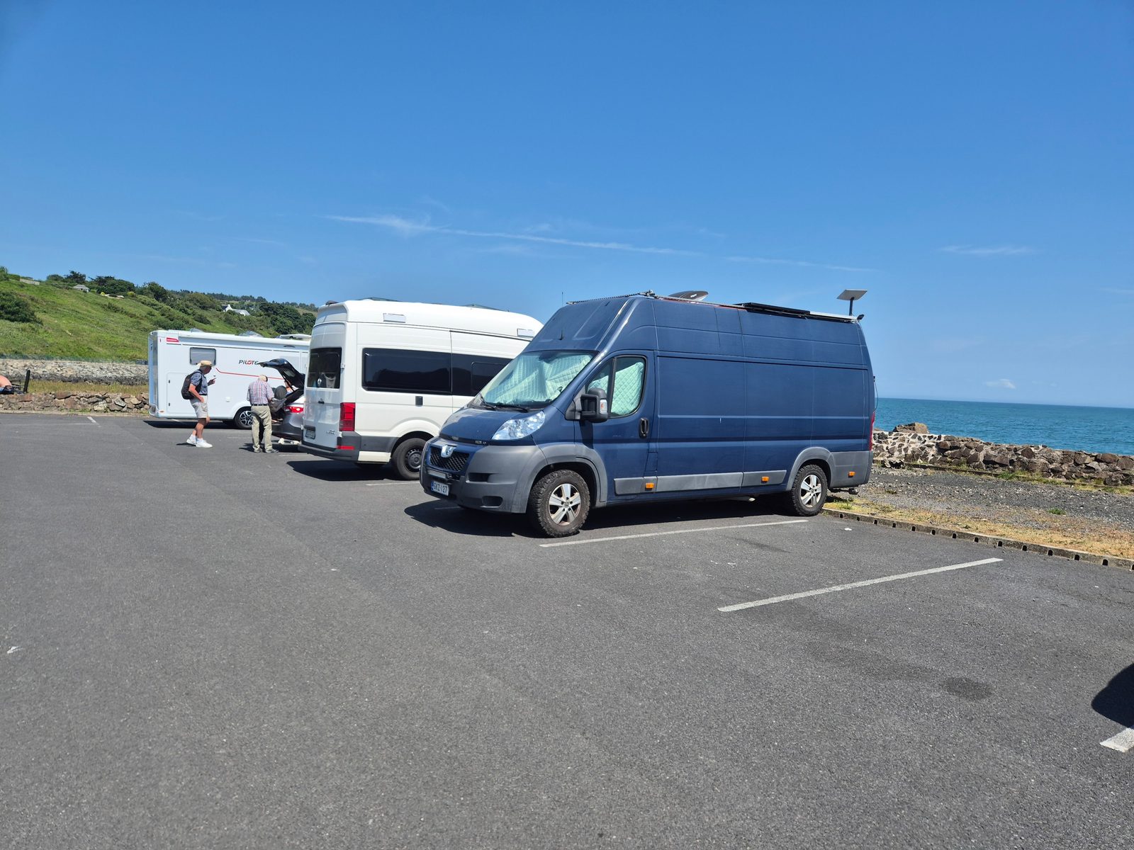 A dark blue Peugeot Boxer panel van with roof solar panels parked beside a white campervan and motorhome in a seaside car park overlooking the sea.