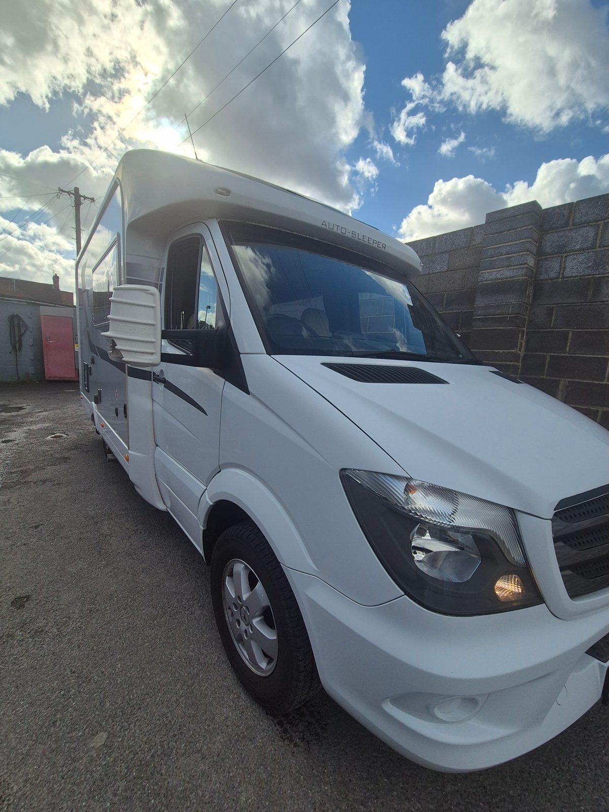 A white Auto-Sleeper Mercedes coachbuilt motorhome parked outside the garage under a blue sky with clouds.