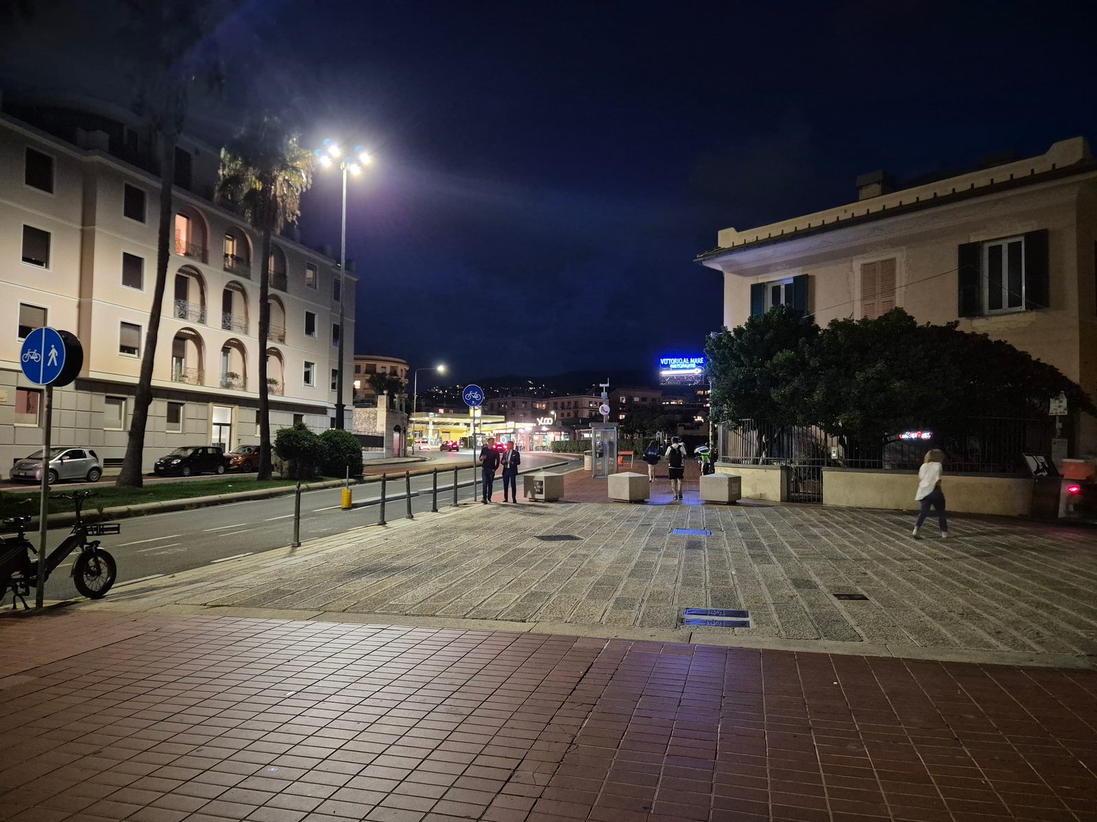 A town square at night with palm trees, apartment buildings and a few people walking.