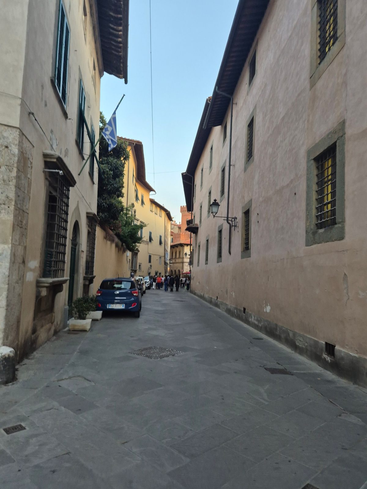A narrow stone-paved street between tall old buildings in an Italian town, with a blue car parked and people walking in the distance.