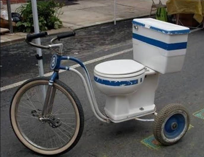 A homemade three-wheeled tricycle made from a white toilet with a cool box cistern, blue handlebars and mismatched wheels, parked on a road.