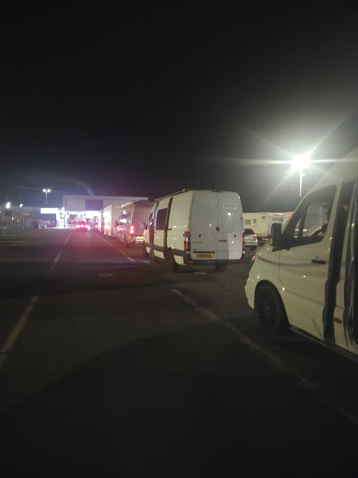 A line of vans and a motorhome queuing at a ferry terminal at night under floodlights.