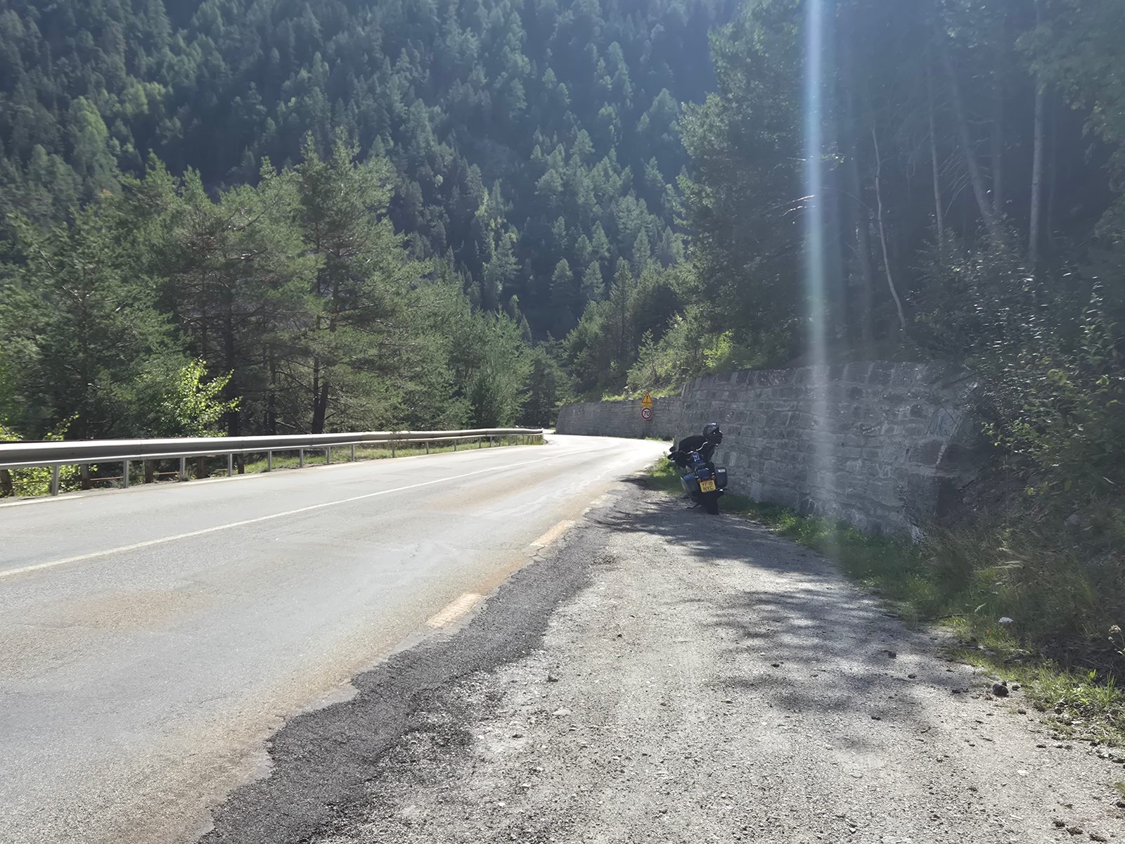 A motorcycle parked by a stone wall on a quiet mountain road surrounded by pine-covered slopes.