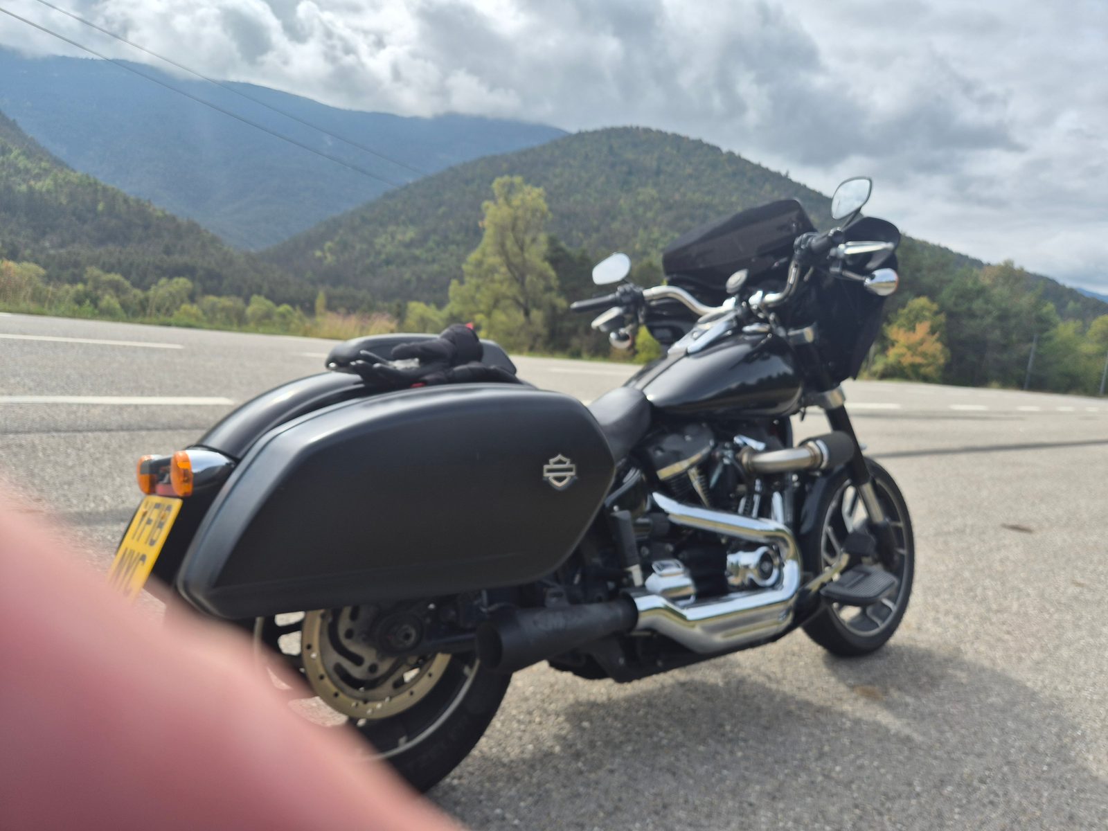 A black Harley-Davidson with panniers parked at the roadside with forested mountains behind.