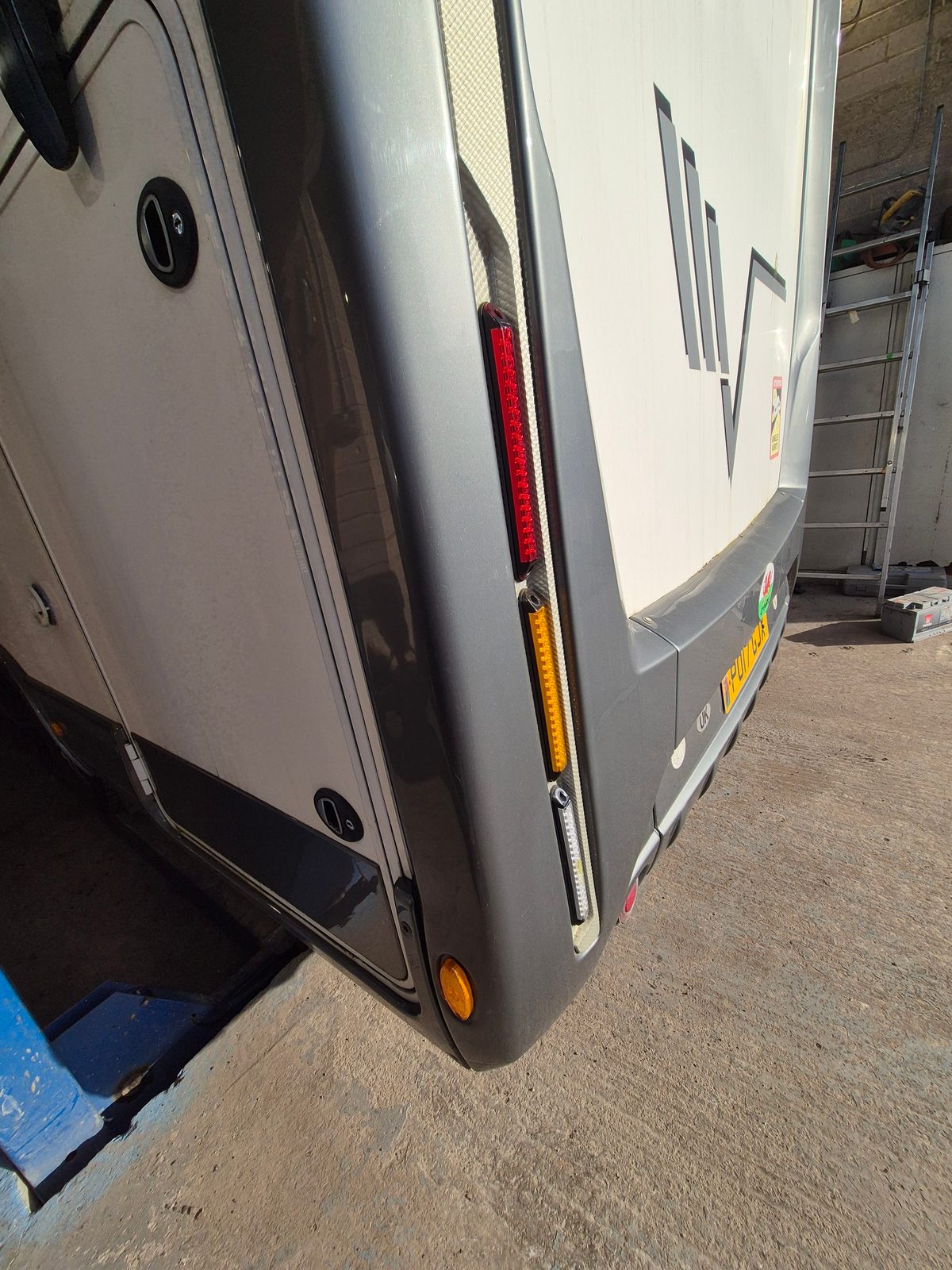 Close-up of the rear nearside corner of a grey and white motorhome, showing the rear light cluster, indicator and bumper, parked by a garage wall with a ladder nearby.