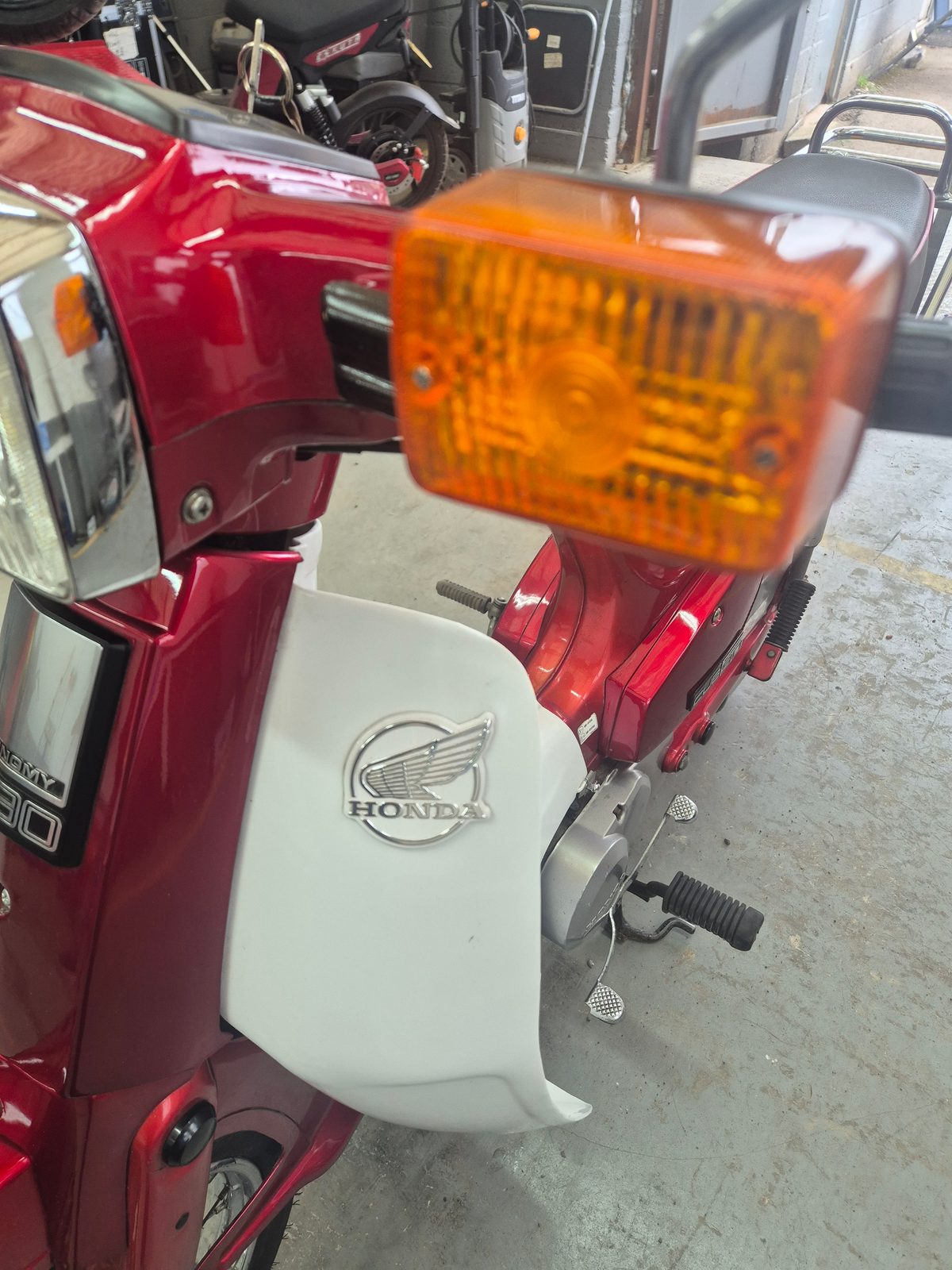 Close-up of a red and white Honda moped showing the front indicator, Honda wing badge and kickstart, with another motorcycle behind on a garage floor.