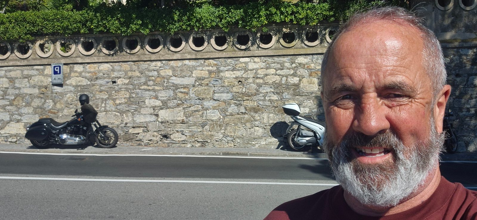 A smiling bearded man taking a selfie beside a stone wall with motorcycles parked along the roadside behind him.