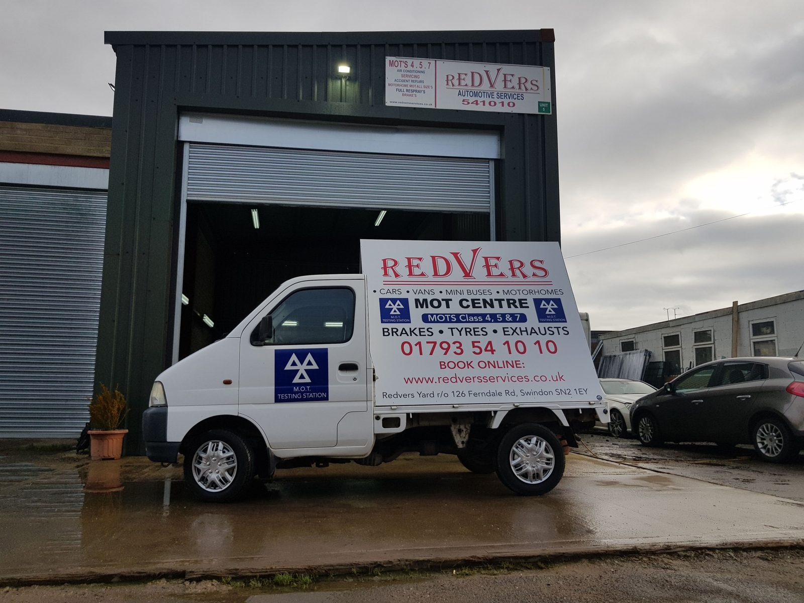 A small white pickup truck displaying a Redvers MOT Centre advertising board, parked in front of an open garage roller door under a grey sky.