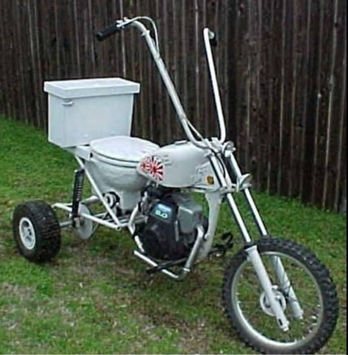 A custom three-wheeled petrol-engined trike with a white toilet and cistern used as the seat, parked on grass in front of a wooden fence.