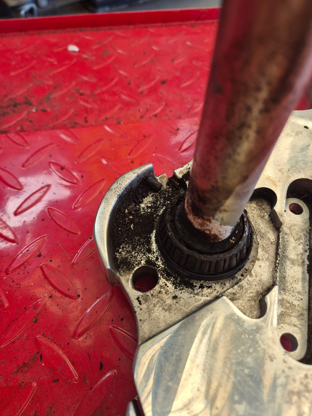 A tapered roller bearing and shaft resting on a red diamond-plate workbench, with dirt and grease around the bearing and a metal bracket alongside.