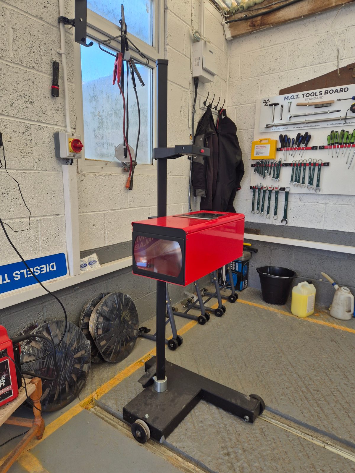 A red headlamp beam tester on a wheeled stand in an MOT bay, with a tool board and spanners on the wall behind.