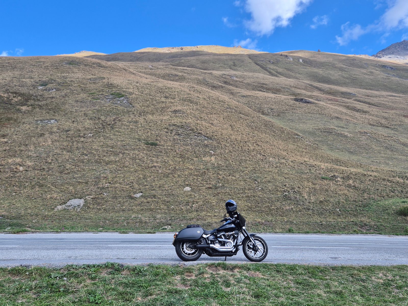 A black Harley-Davidson motorcycle parked on a mountain road below a grassy hillside under a blue sky.
