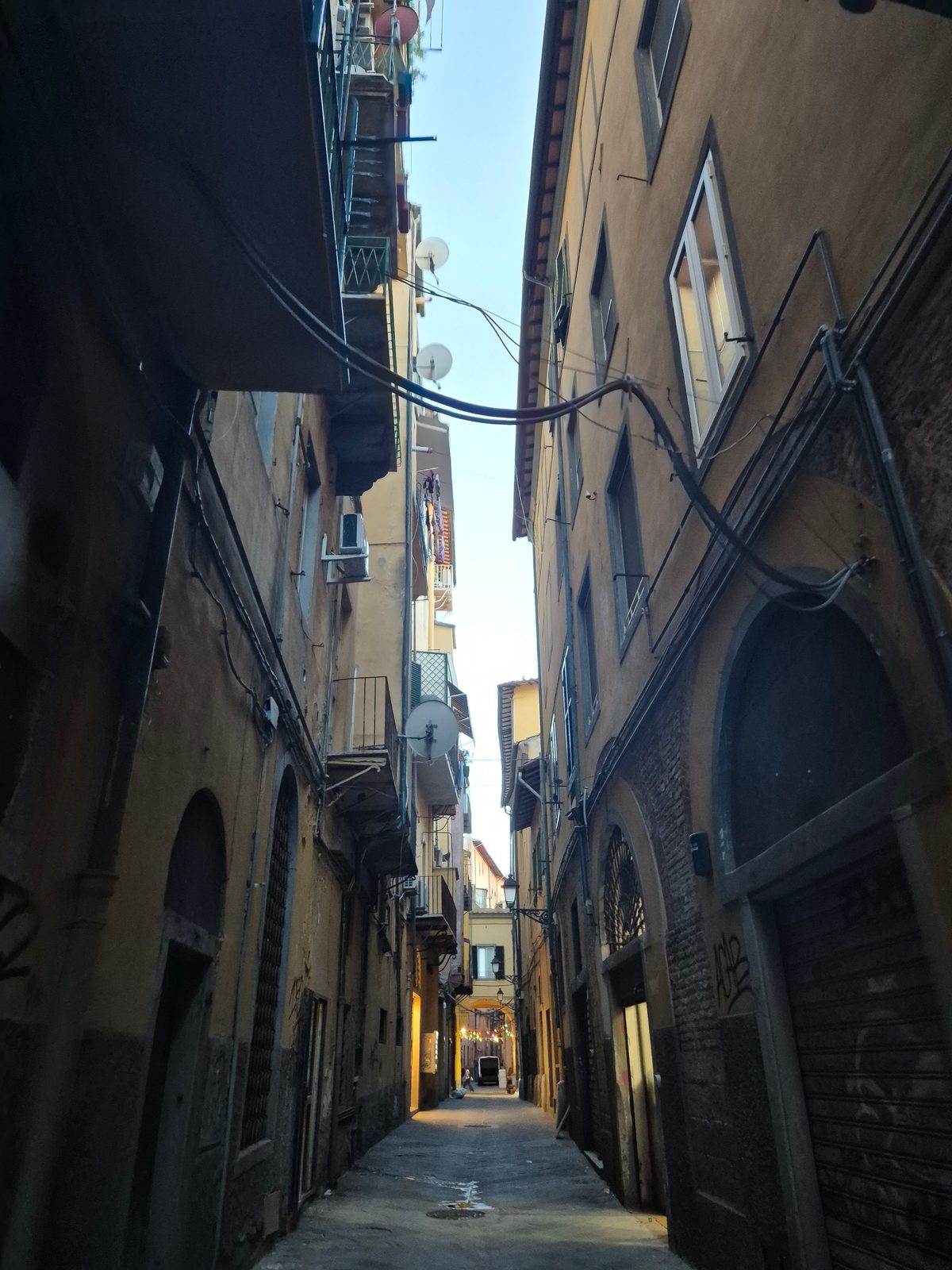 A narrow Italian alley between tall old buildings with shuttered windows, balconies and overhead cables.