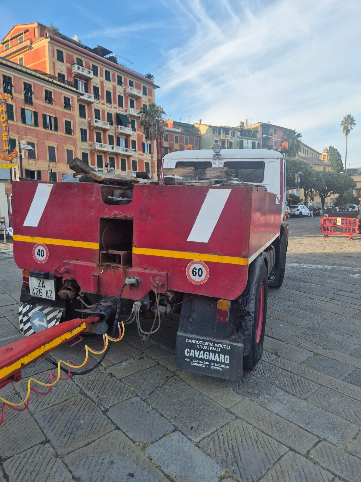 An old red Italian tipper truck parked on a stone piazza in front of colourful seafront buildings.