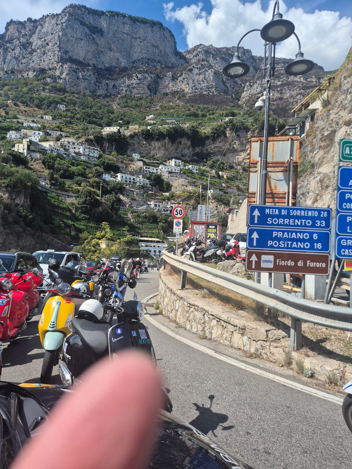 A row of parked Vespa scooters beside a coastal road in Italy with signs for Sorrento and Positano and mountains behind.