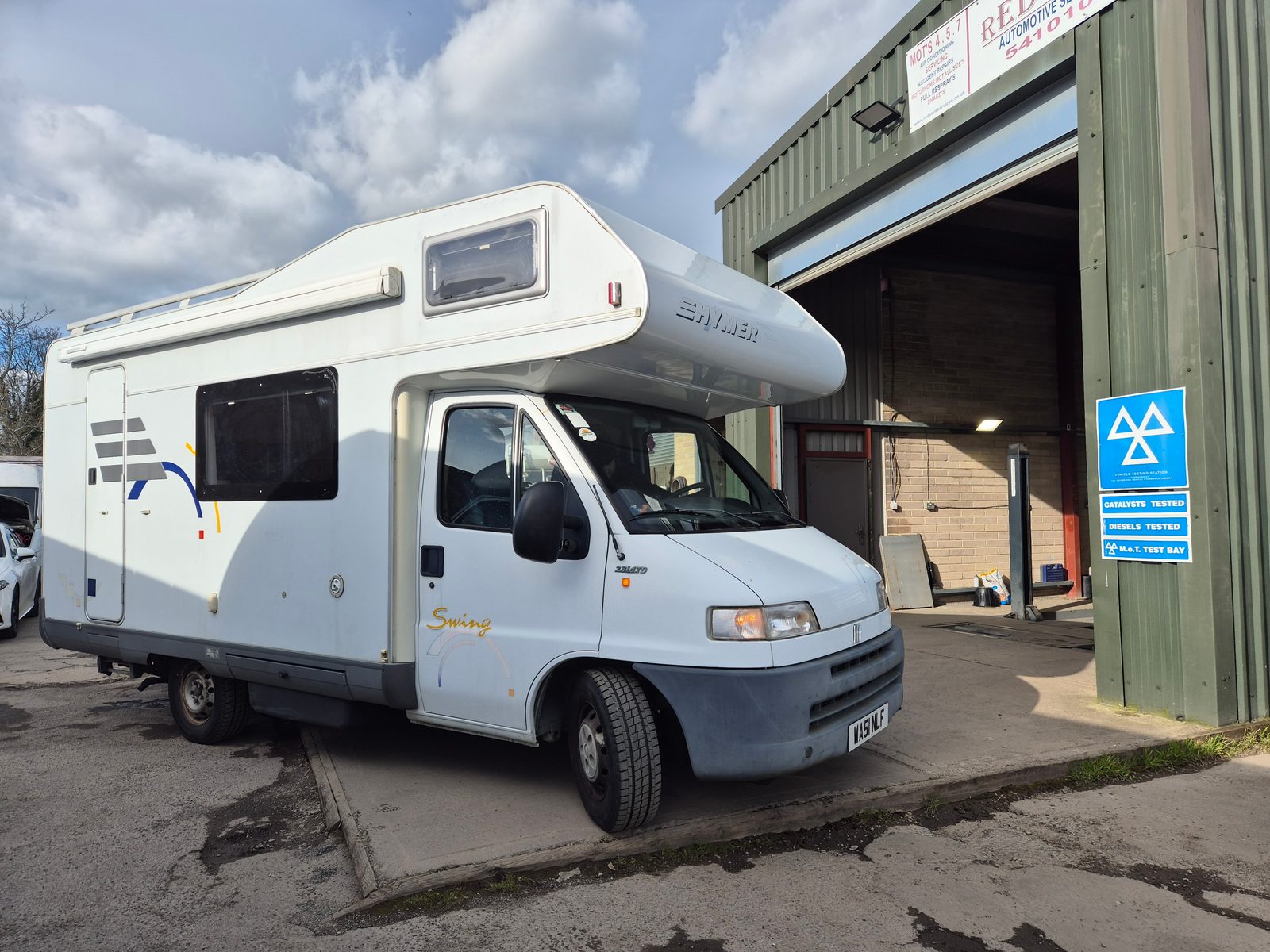 A white Hymer Swing coachbuilt motorhome on a Fiat base parked beside the green garage entrance, with an MOT test bay sign on the wall.