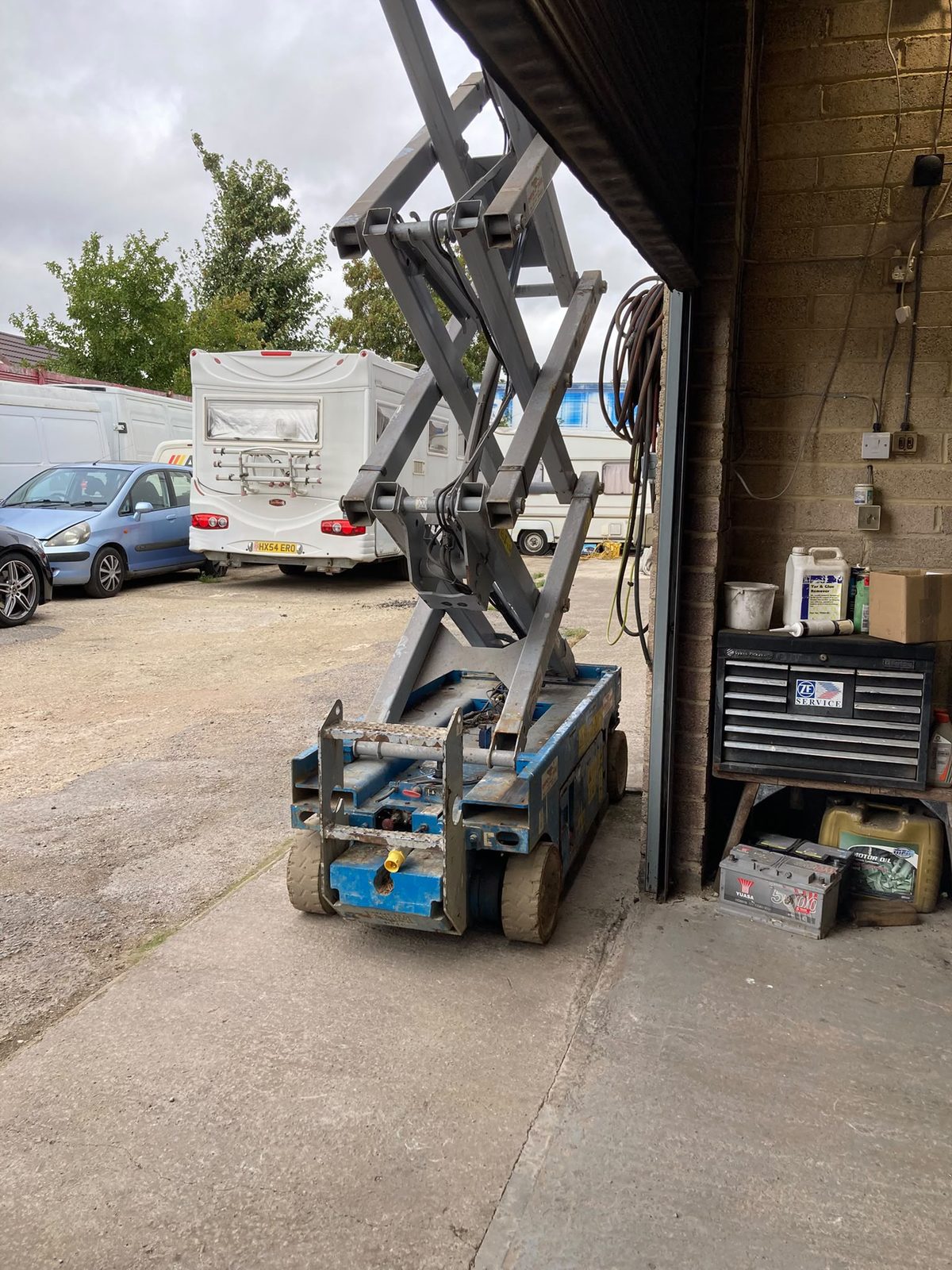 A blue scissor lift parked by the garage entrance with motorhomes and a car in the yard behind.