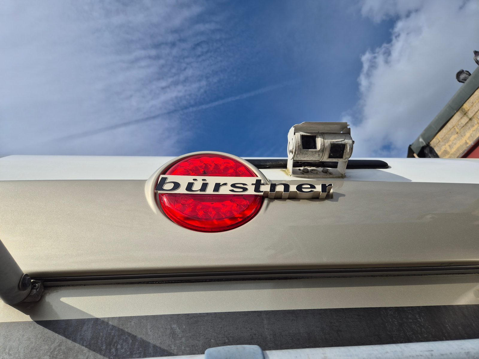 Close-up of a Bürstner motorhome's rear panel with a round red LED light, Bürstner badge and a reversing camera against a blue sky.